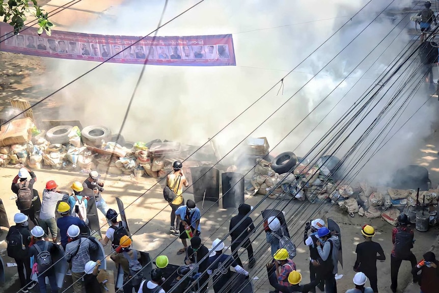 Protesters wearing construction hard hats stand back from a makeshift barricade as a thick cloud of smoke fills the area.