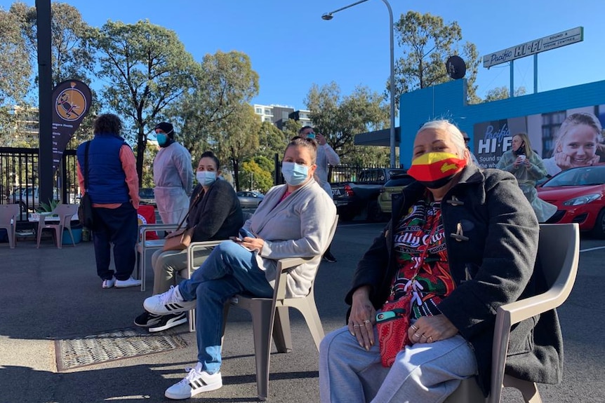 Three Indigenous women sit on chairs in an outdoor COVID vaccination clinic.