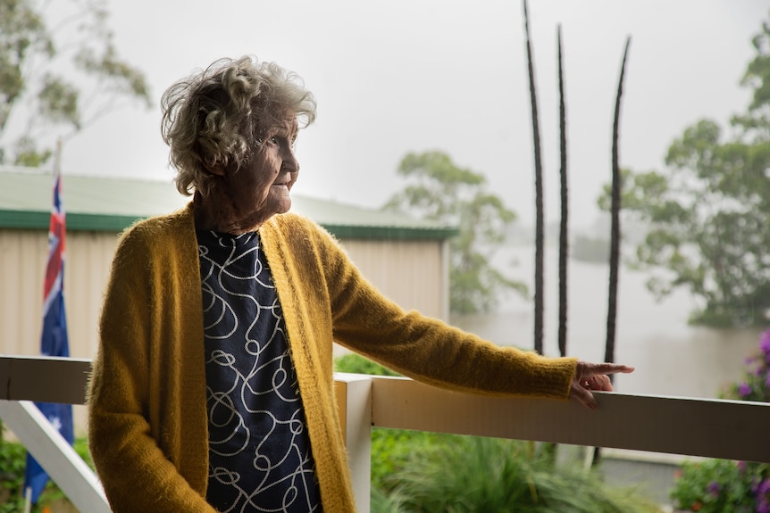 An elderly woman stands on a deck overlooking floodwaters in her backyard.