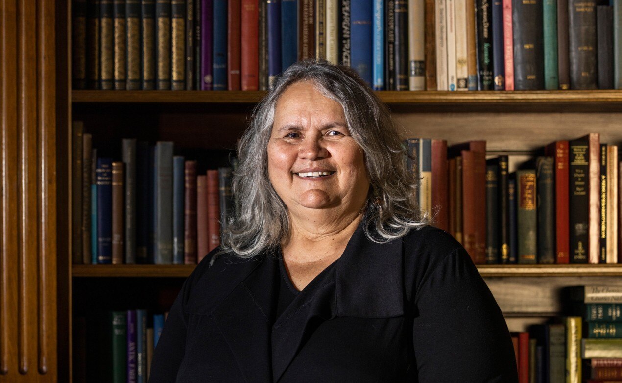 An Indigenous woman with shoulder-length grey hair poses for a portrait in front of shelves of books, smiling warmly.