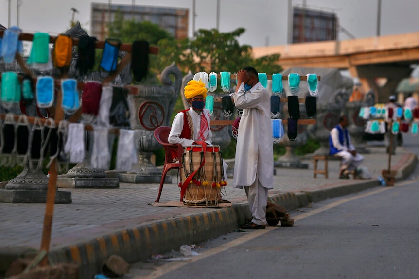 On an overcast day, you view a brightly-dressed man in a red plastic chair amid rows of face masks for sale.