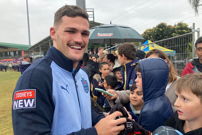 Nathan Cleary in a NSW Blues jacket smiles as he signs autographs.