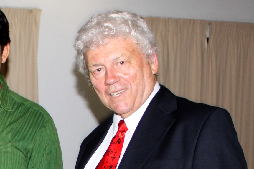 A man wearing a suit with grey hair, turns his head to smile at the camera.