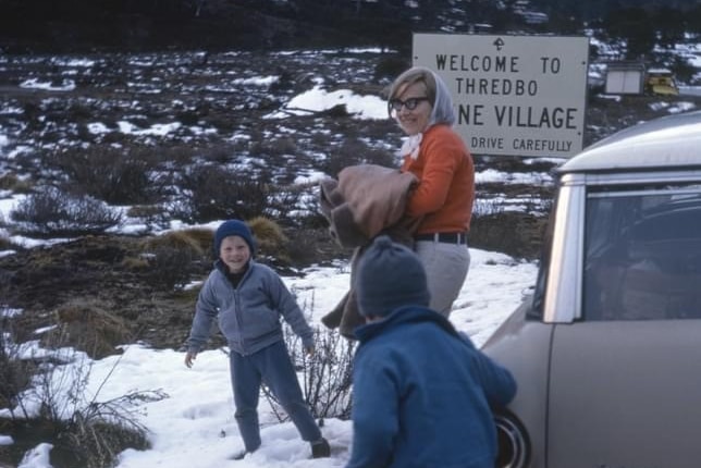 A 1960s mother watches as two young children play in the snow in front of a car with a Thredbo sign behind them 