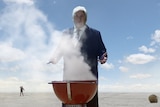 A white-haired man in a suit stands over a small smoking barbecue in a desert.