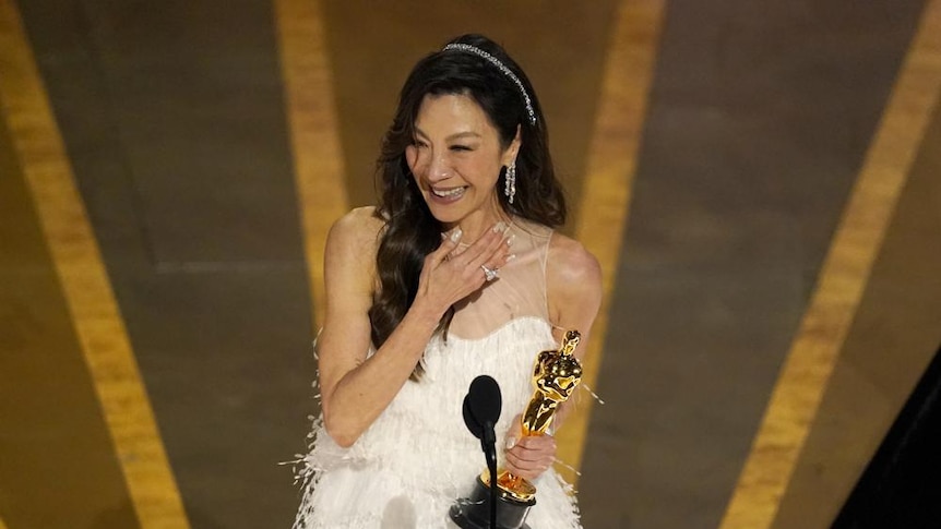 Actor Michelle Yeoh wears white dress, smiling from stage, hand on chest, as she accepts an Oscar award. 