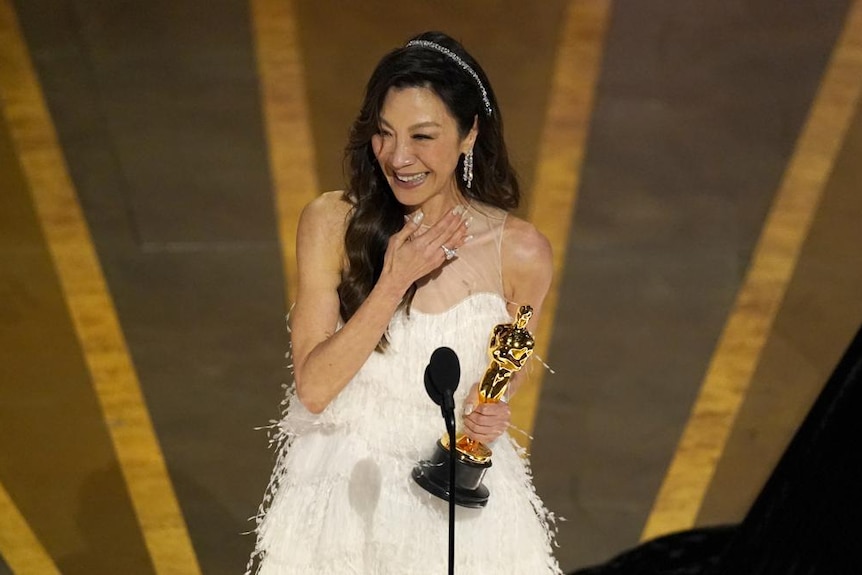 Actor Michelle Yeoh wears white dress, smiling from stage, hand on chest, as she accepts an Oscar award. 