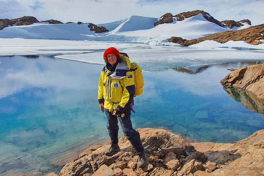 A woman wearing a yellow and black jacket and red beanie stands in front of a clear blue lake snow behind.