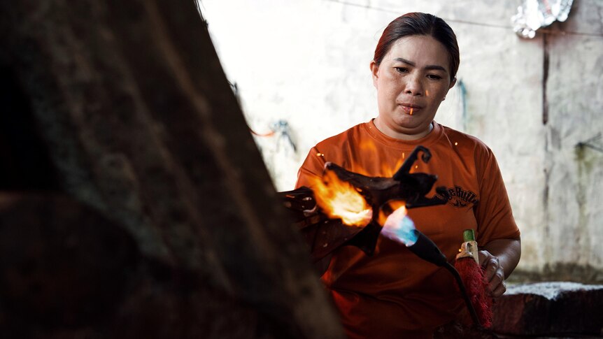 A woman wearing an orange shirt holds a blowtorch to a a bat