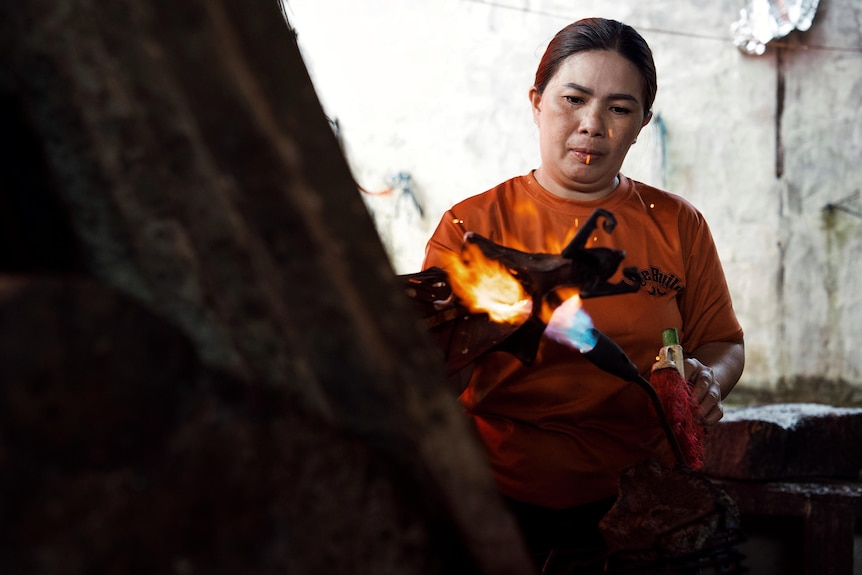 A woman wearing an orange shirt holds a blowtorch to a a bat