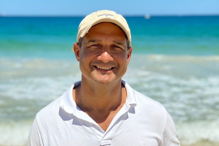 Un hombre con camisa blanca y gorra parado en una playa y sonriendo ante el cañón de la cámara.