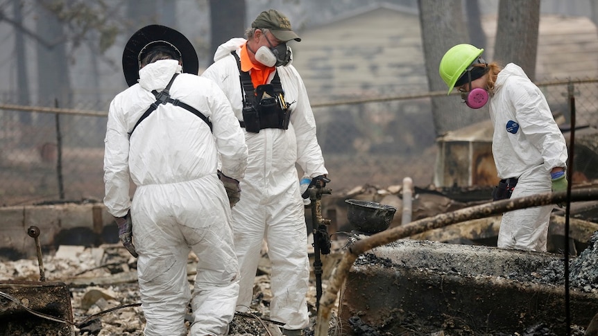 Three people wearing masks and protective gear search through ashes and burnt items inside the site of a destroyed house.