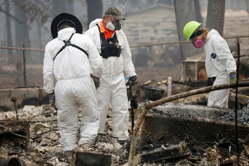 Three people wearing masks and protective gear search through ashes and burnt items inside the site of a destroyed house.