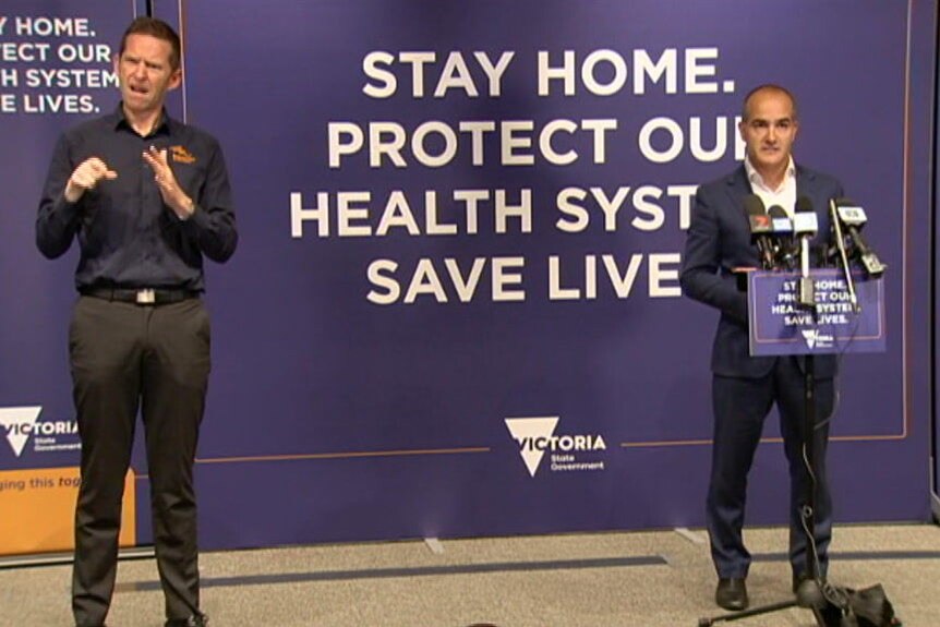 An AUSLAN interpreter and James Merlino stand at a distance from one another in front of a purple 'stay home' sign.