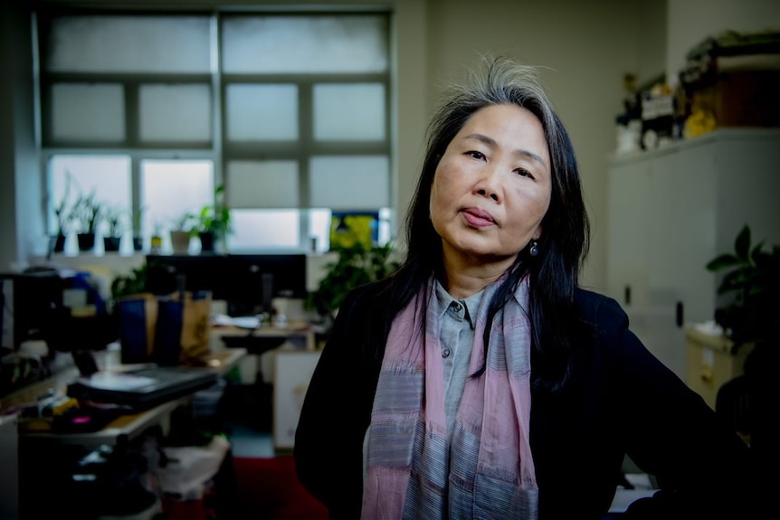 A woman wearing a dark cardigan stands in an office, with her hand on her hip.