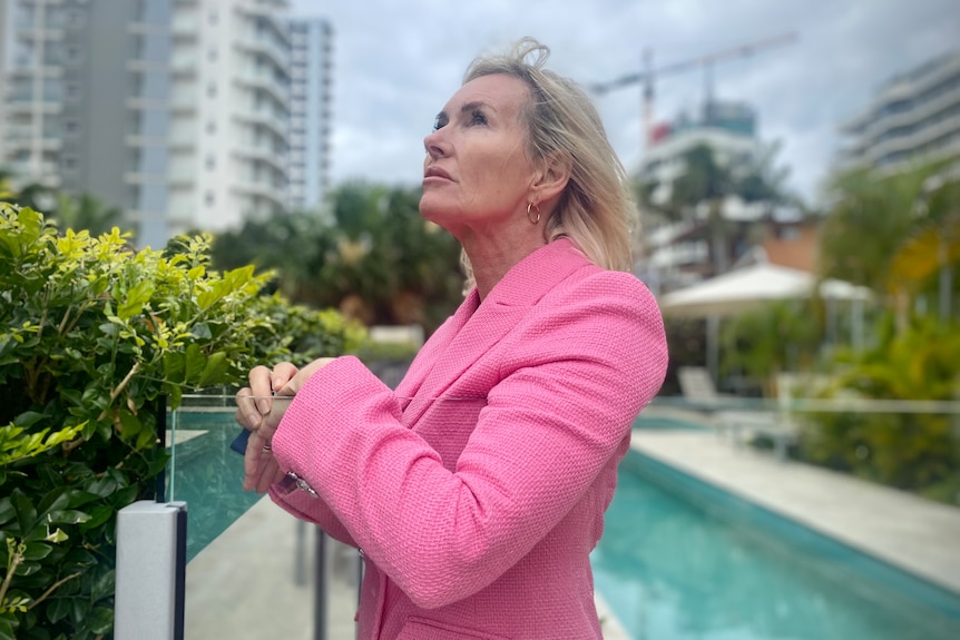 A woman in a pink jacket looks at high-rise apartment towers while standing by a pool.