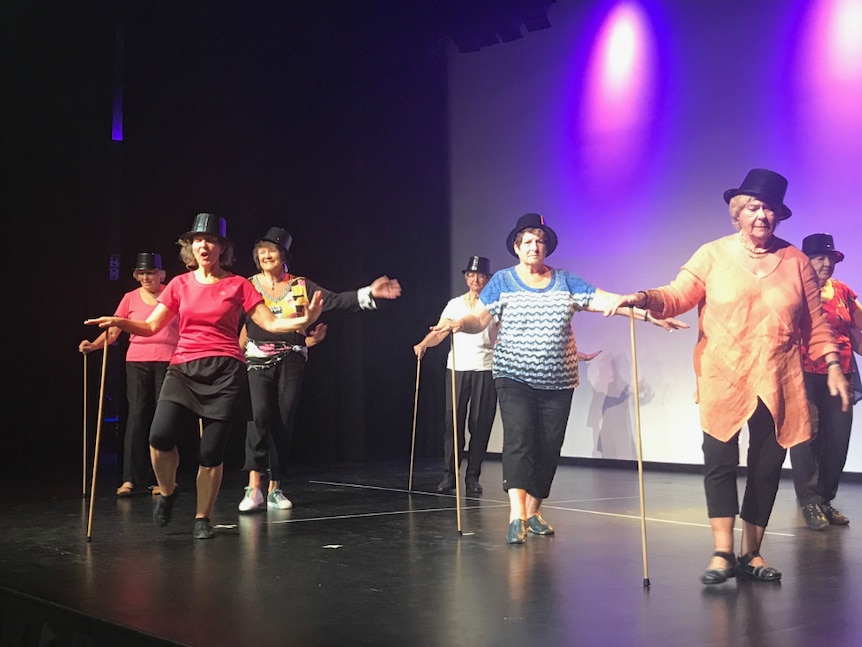 Senior women dancing on stage in plain clothes with top hat, cane stick, lighting in the background, in dance moves.