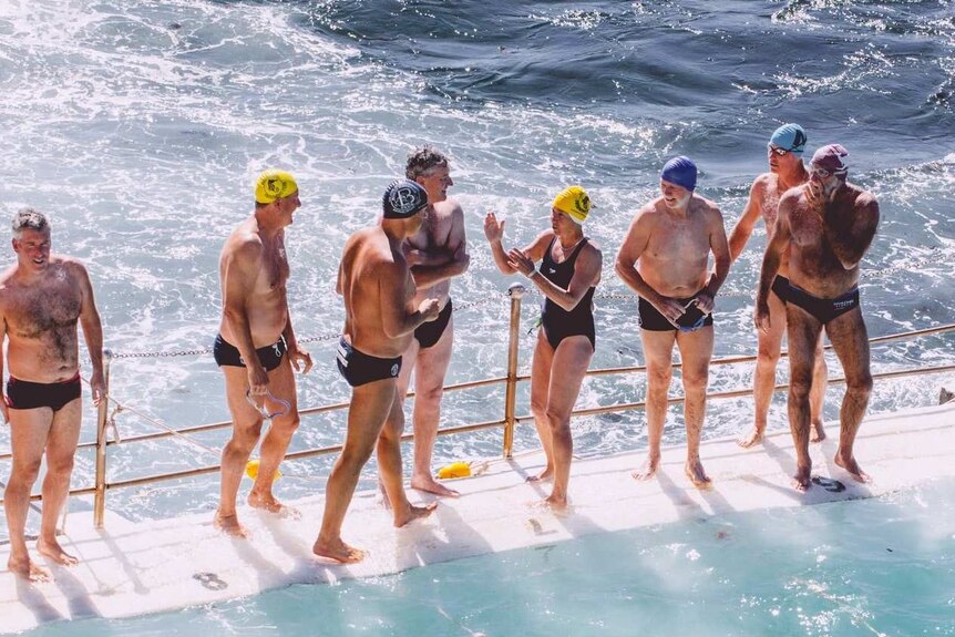 Swimmers of mixed ages stand at the edge of an ocean pool, about to race.
