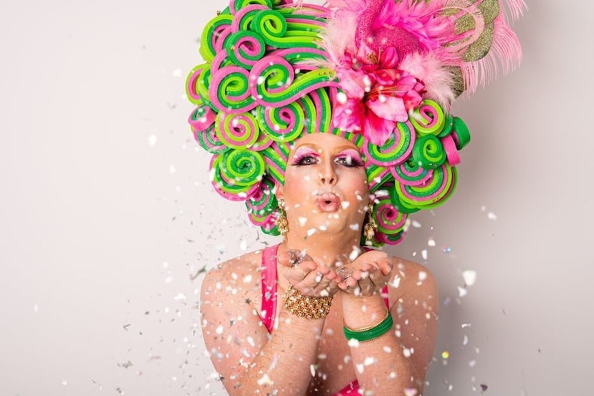 A drag queen with a green and pick hat blowing confetti 