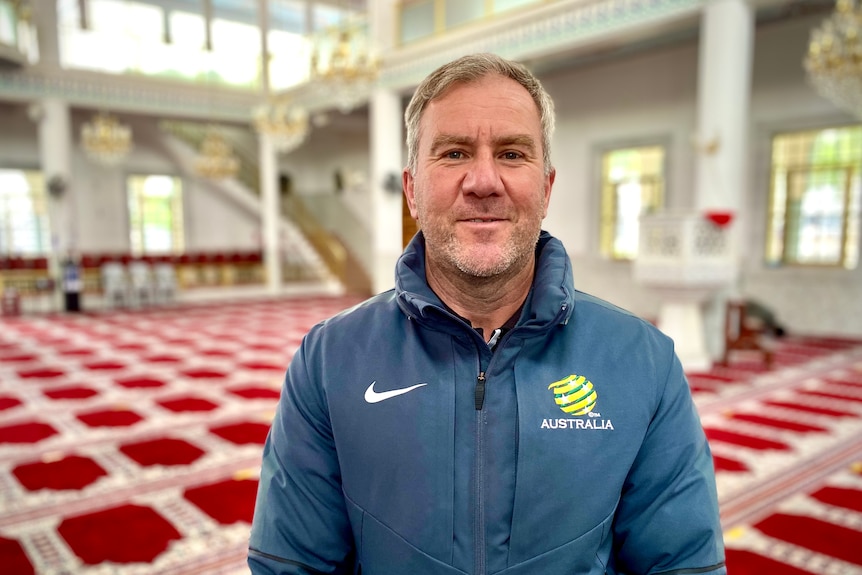 A man wearing a jacket smiles into the camera while standing in a large carpeted room.