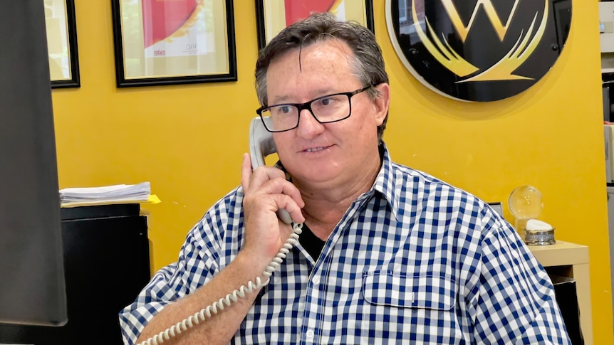 Man wearing a checked shirt sits at a desk while speaking on the phone.