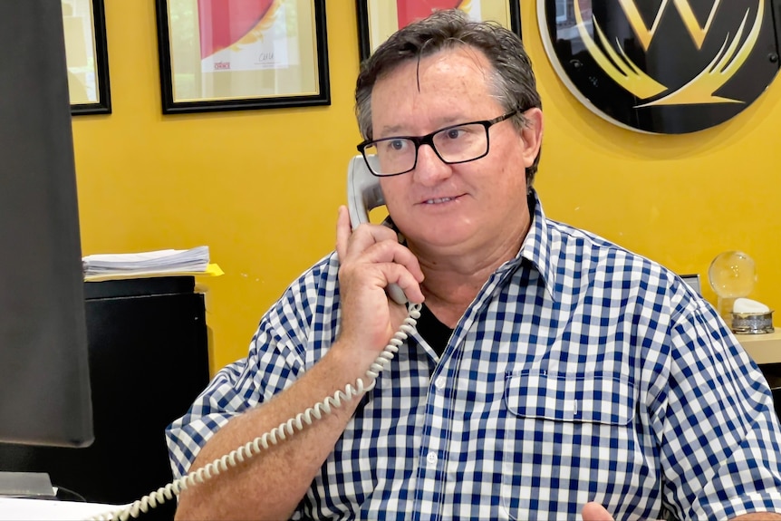 Man wearing a checked shirt sits at a desk, speaking on the phone.