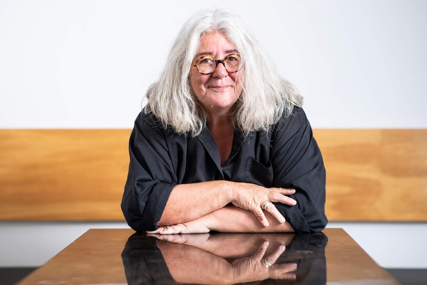 Woman with shoulder-length white hair sitting at table, arms folded, wearing round tortoiseshell-framed glasses and black shirt.