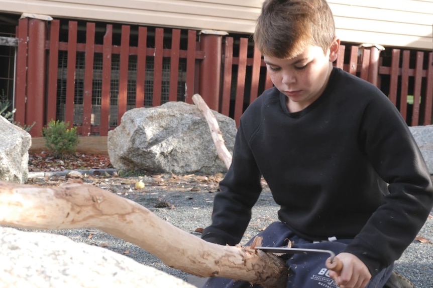 A young boy kneels with a didgeridoo resting on a rock and between his legs as he uses a tool to carve it's outside