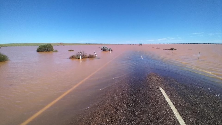 Flat straight road covered by red muddy water under a brilliant blue sky.