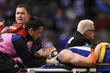 An AFL player in a neck brace lies on a stretcher with eyes closed as he is attended to.
