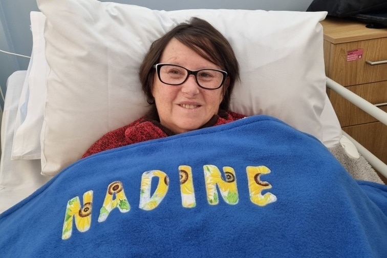 A woman wearing glasses in bed with a blue blanket across her with the words "Nadine"