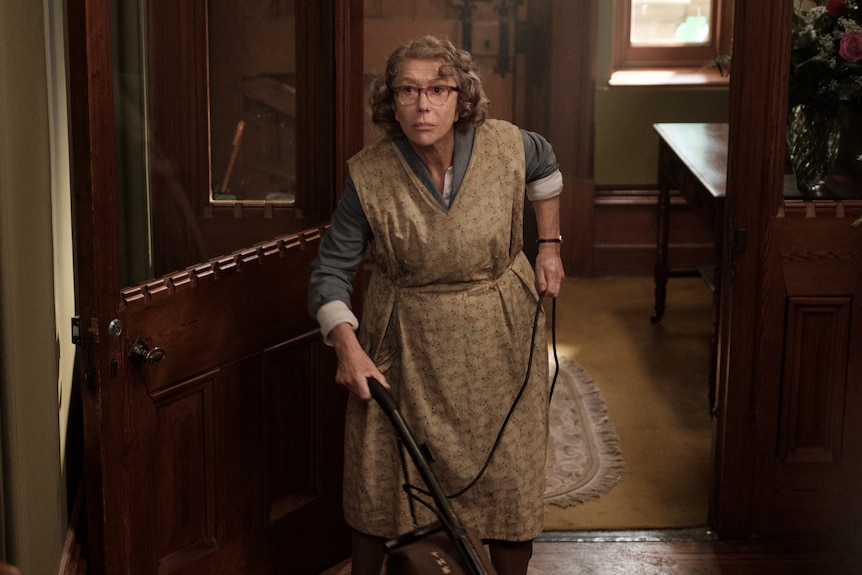 60-something year old white woman with grey hair and glasses wears 60s cleaning dress and vacuums hallway