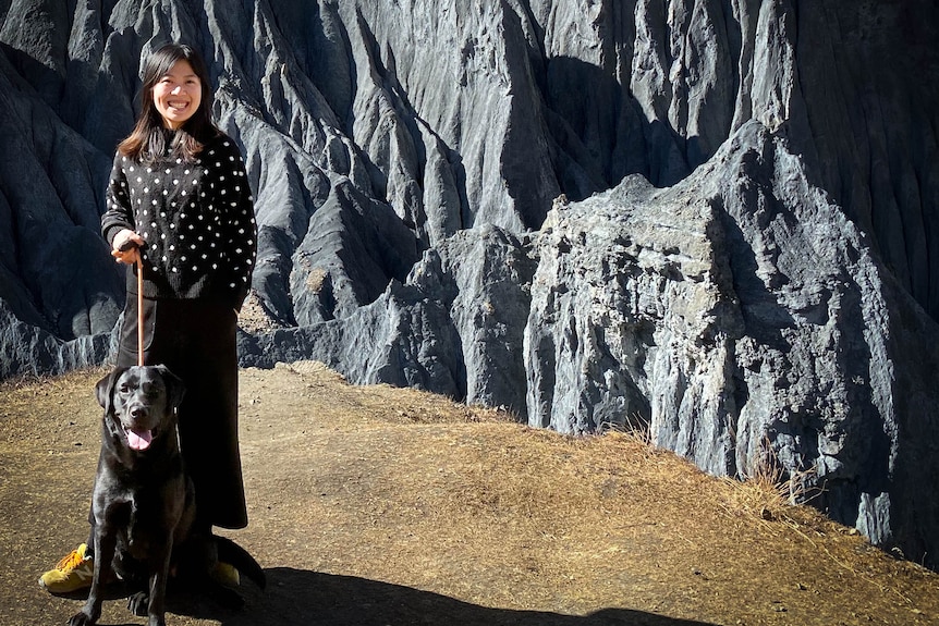 woman in spotty jumper stands in front of rocky mountain range with black dog on lead