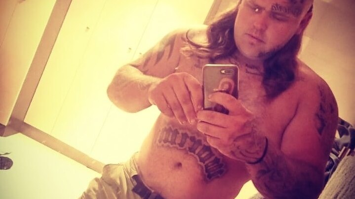 A shirtless man with a pronounced mullet and many tattoos, including on his face, takes a selfie in the mirror.
