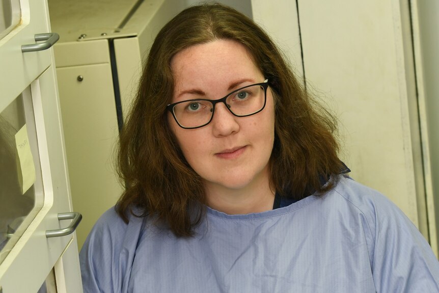 A woman wearing medical scrubs looks seriously at the camera.