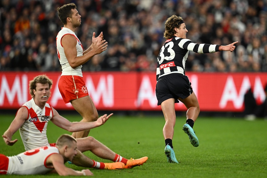 An Aussie rules player wearing black and white celebrates in front of opponents in red and white at packed stadium