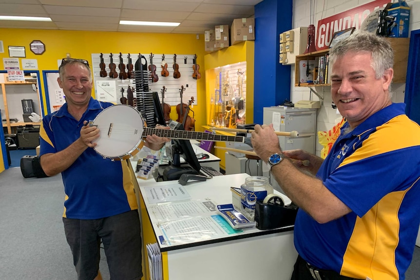 A mid shot of two men standing at a counter in a music shop with a banjo stretched out before them.