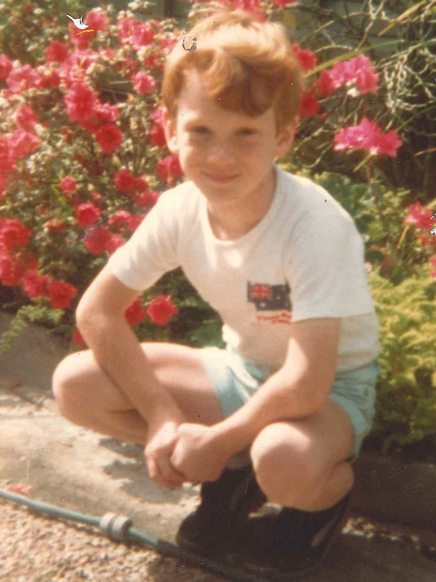 A young boy squats down next to some flowers, smiles at the camera.