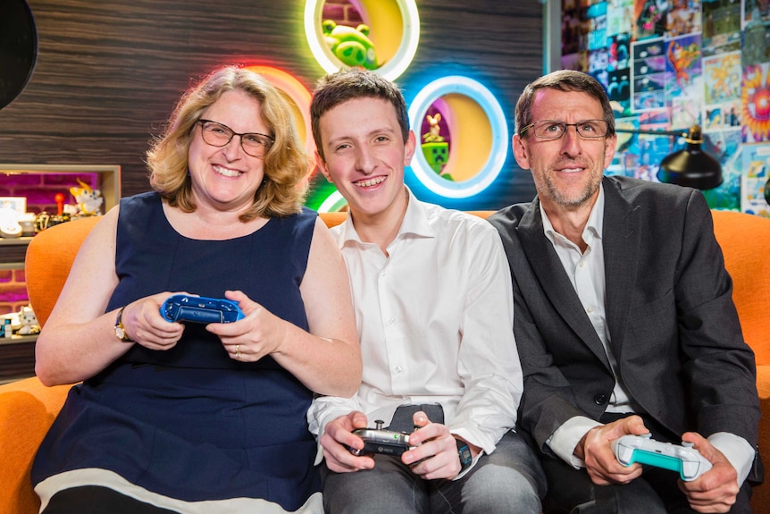 A woman, a young man and a man sit on a lounge holding electronic game controllers.