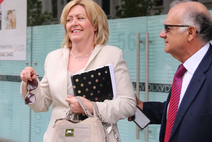 Lisa Scaffidi smiles holding a notepad, handbag and pair of sunglasses outdoors with her husband Joe alongside her.