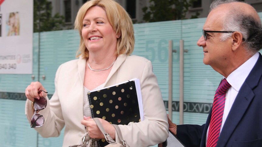 Lisa Scaffidi smiles holding a notepad, handbag and pair of sunglasses outdoors with her husband Joe alongside her.