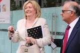 Lisa Scaffidi smiles holding a notepad, handbag and pair of sunglasses outdoors with her husband Joe alongside her.