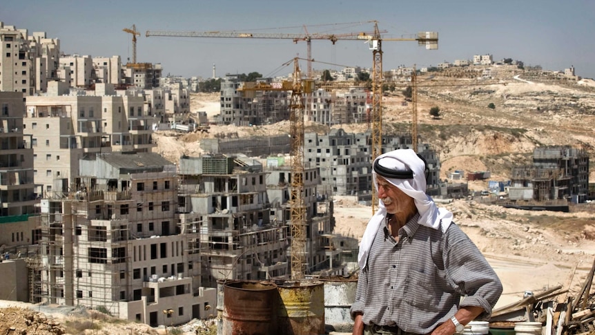 A Palestinian man stands next to his house in front of construction sites and cranes.