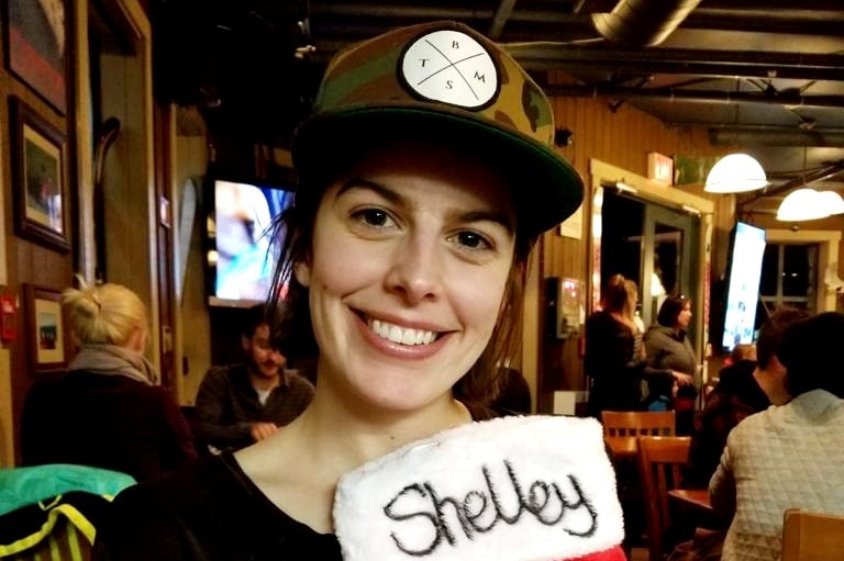 A woman wearing a trucker cap and holding a Christmas stocking with the word 'Shelley' on it stands in a restaurant.