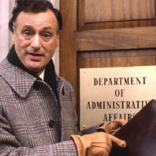 British actor Paul Eddington holds a briefcase and stands in front of a door marked 'Department of Administrative Affairs'