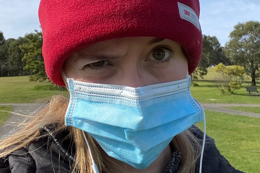 A woman in a red beanie and blue surgical mask raises an eyebrow in a selfie taken in a park under blue skies.