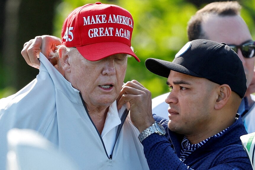 A man wearing a black cap fixes the collar of Trump's white jumper.