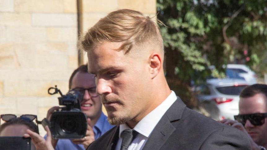 A man in a suit looks at the ground as a crowd film him in the background.