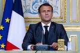 Emmanuel Macron sits in on a desk in front of an ornate panelled wall with gold accents, next to French and EU flags.
