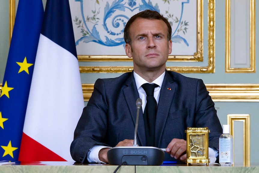 Emmanuel Macron sits in on a desk in front of an ornate panelled wall with gold accents, next to French and EU flags.